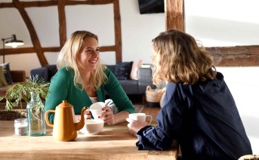 Zwei Frauen sitzen sich l&auml;chelnd an einem Holztisch gegen&uuml;ber und unterhalten sich bei Tee oder Kaffee. Im Hintergrund ist ein rustikaler Raum mit dunklem Fachwerk erkennbar.