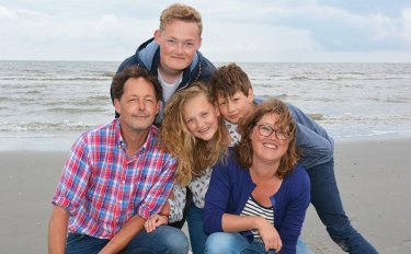 Die Familie Tuinier Hofman posiert l&auml;chelnd f&uuml;r ein Gruppenfoto an einem grauen Sandstrand. Im Hintergrund sind die Wellen des Meeres unter einem bew&ouml;lkten Himmel zu sehen.
