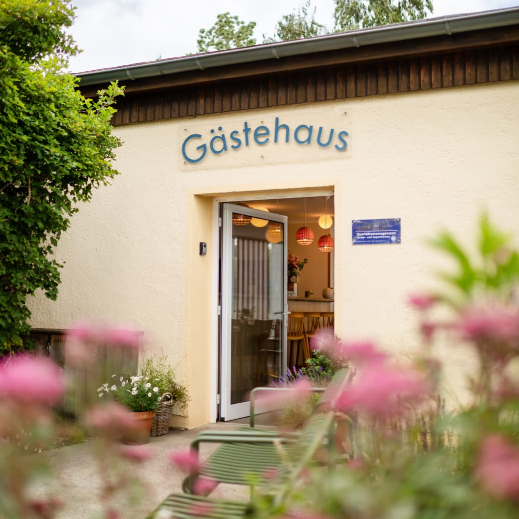 Außenansicht des Osteegästehauses mit geöffneter Tür, Blumen im Vordergrund und Blick in den freundlich beleuchteten Innenraum.